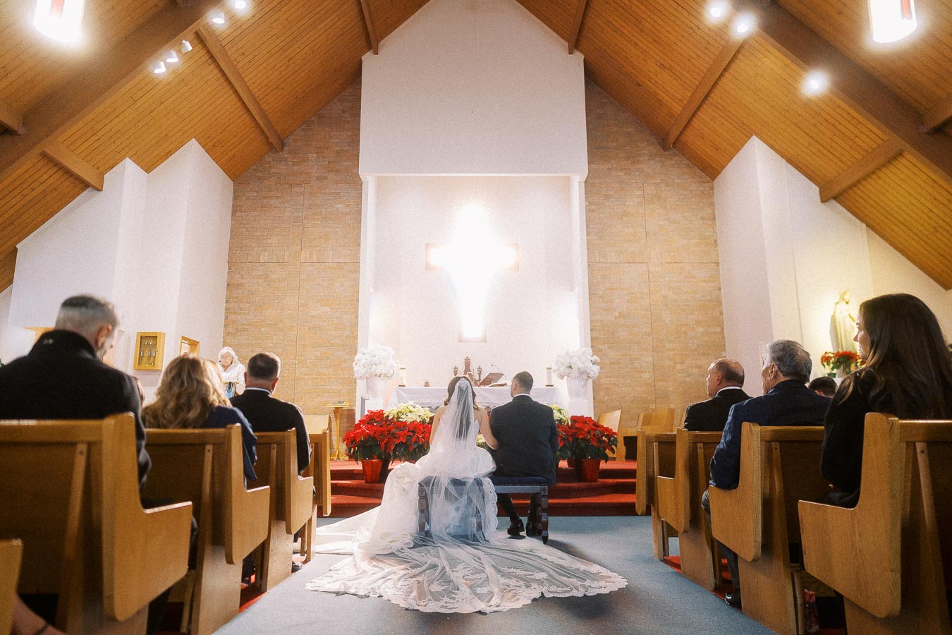 A bride and groom kneel at the altar in a beautifully lit church, surrounded by family and friends seated in wooden pews. The bride's long, lace veil is elegantly spread on the floor, and festive red poinsettias adorn the altar area.