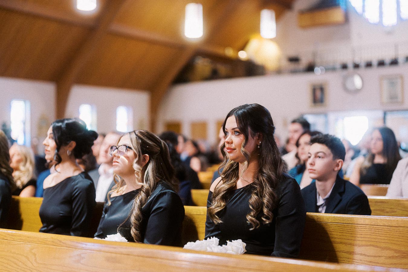 A group of people sitting attentively in a church pew during a ceremony, dressed in formal attire with sunlight filtering through the stained glass windows in the background.