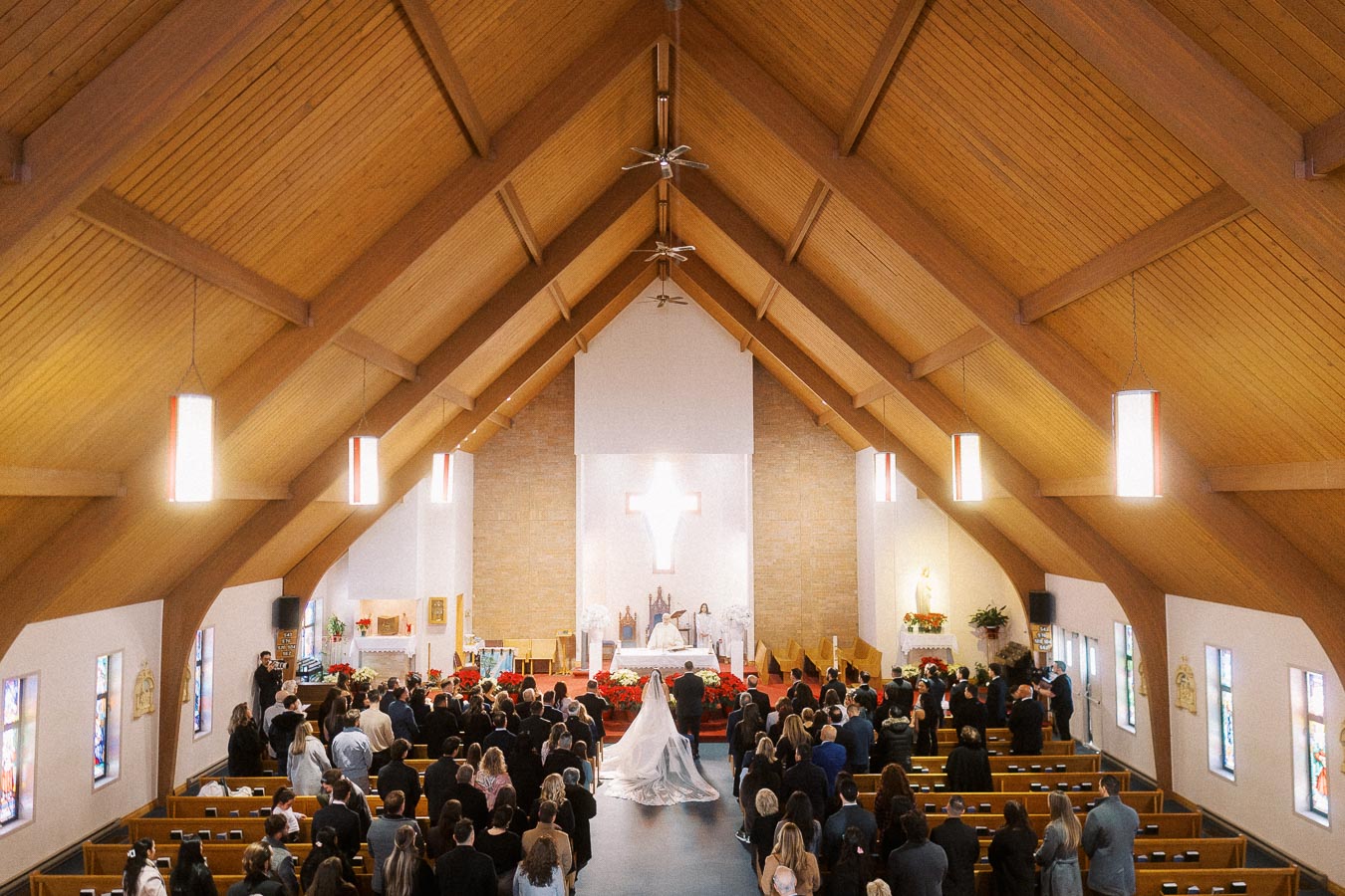 A wedding ceremony taking place inside a beautifully adorned church with a wooden ceiling, stained glass windows, and a crowd of guests seated in pews facing the altar, where a bride in a flowing white gown walks down the aisle.