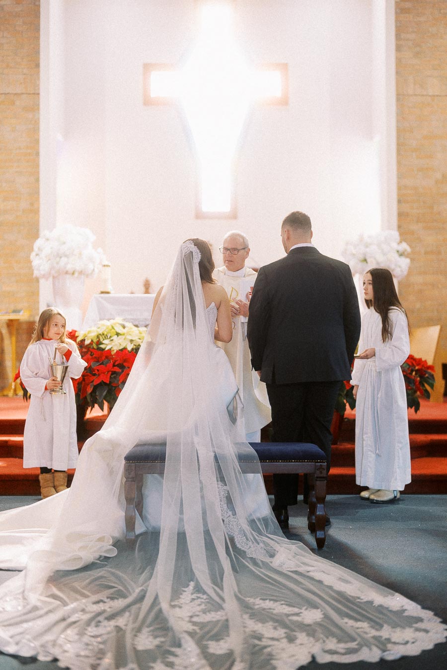A bride in a white gown with a long lace train and a groom in a black suit kneel at the altar during a church wedding ceremony, with a priest officiating and two young altar boys assisting, surrounded by floral arrangements and a large illuminated cross in the background.