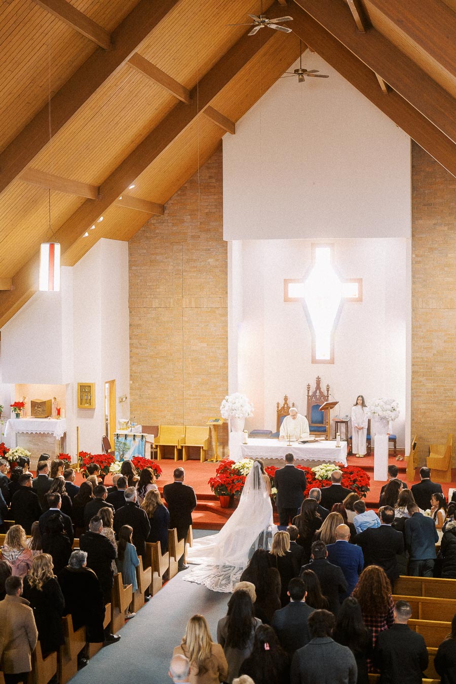 A wedding ceremony taking place in a beautifully decorated church with wooden ceilings, featuring a bride in a long veil walking down the aisle, and guests seated in pews, attentively observing.