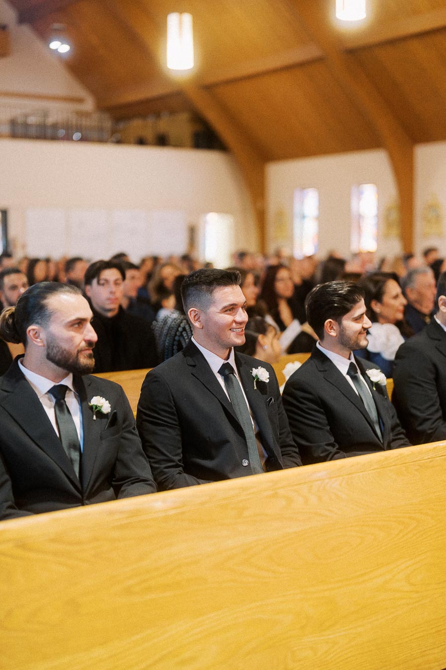 Three groomsmen wearing black suits and boutonnieres, sitting in a church pew, surrounded by guests during a wedding ceremony.