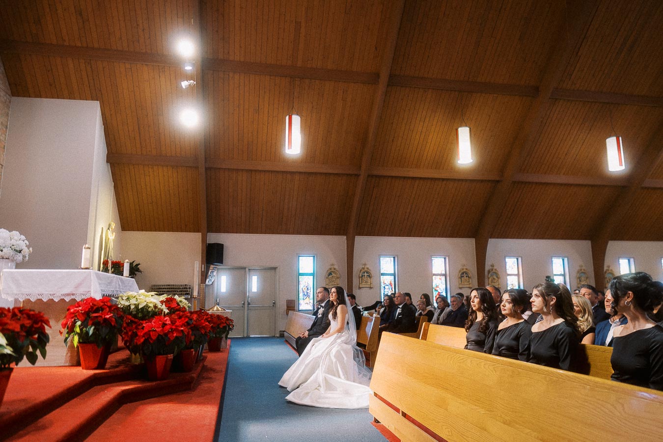 A bride and groom seated at the front of a church during their wedding ceremony, with bridesmaids and guests seated in wooden pews. The church is decorated with red and white poinsettias near the altar, and soft lighting illuminates the wooden ceiling.