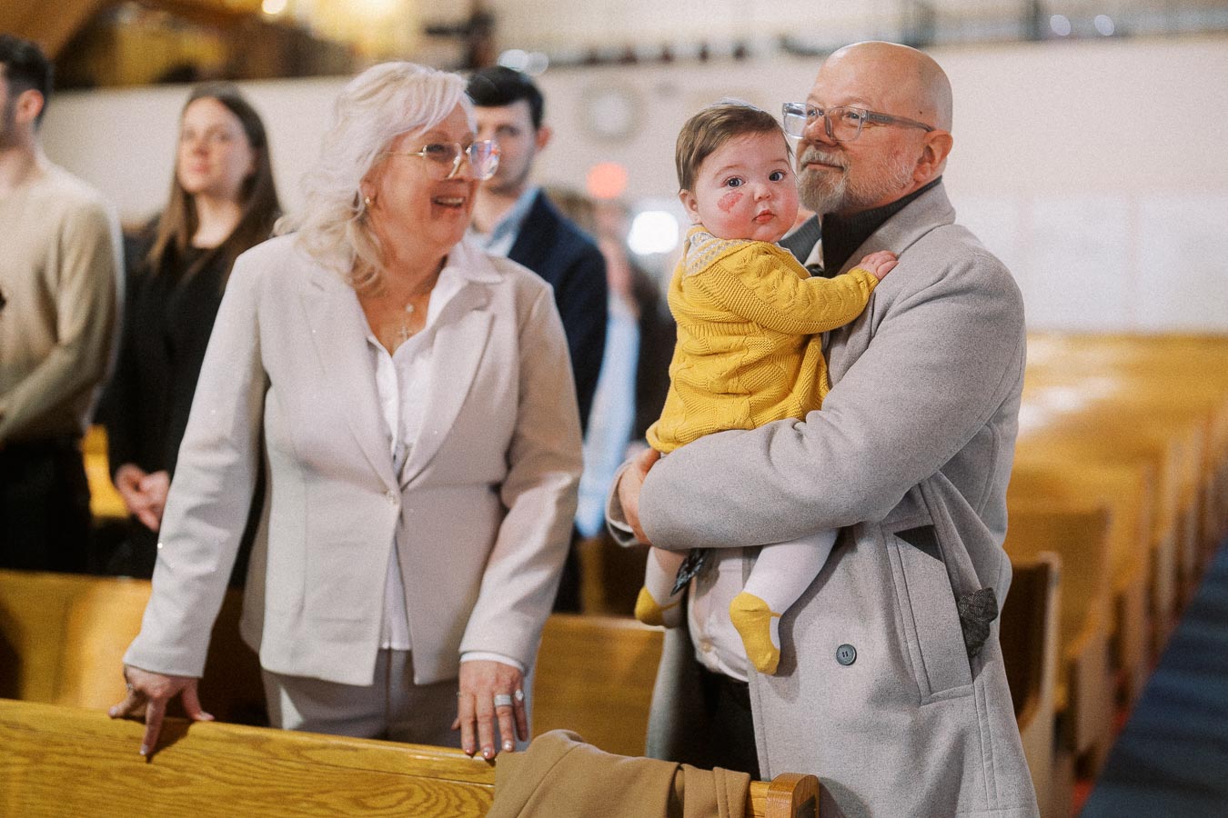 Elderly couple smiling while holding a baby in a church setting, dressed in formal attire.