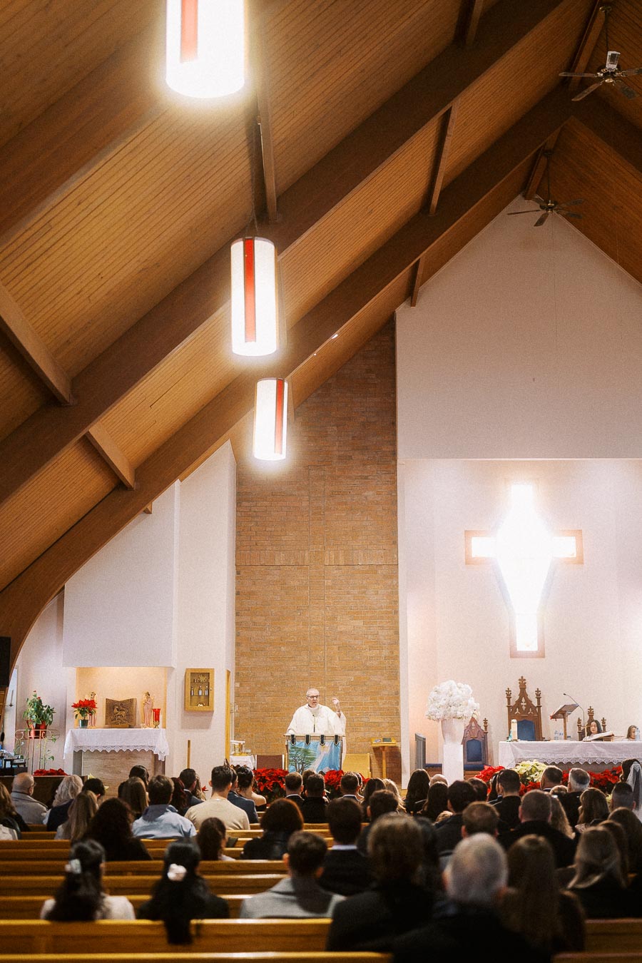 Congregation gathered inside a wooden-ceiling church with priest delivering sermon at altar; illuminated cross in background.