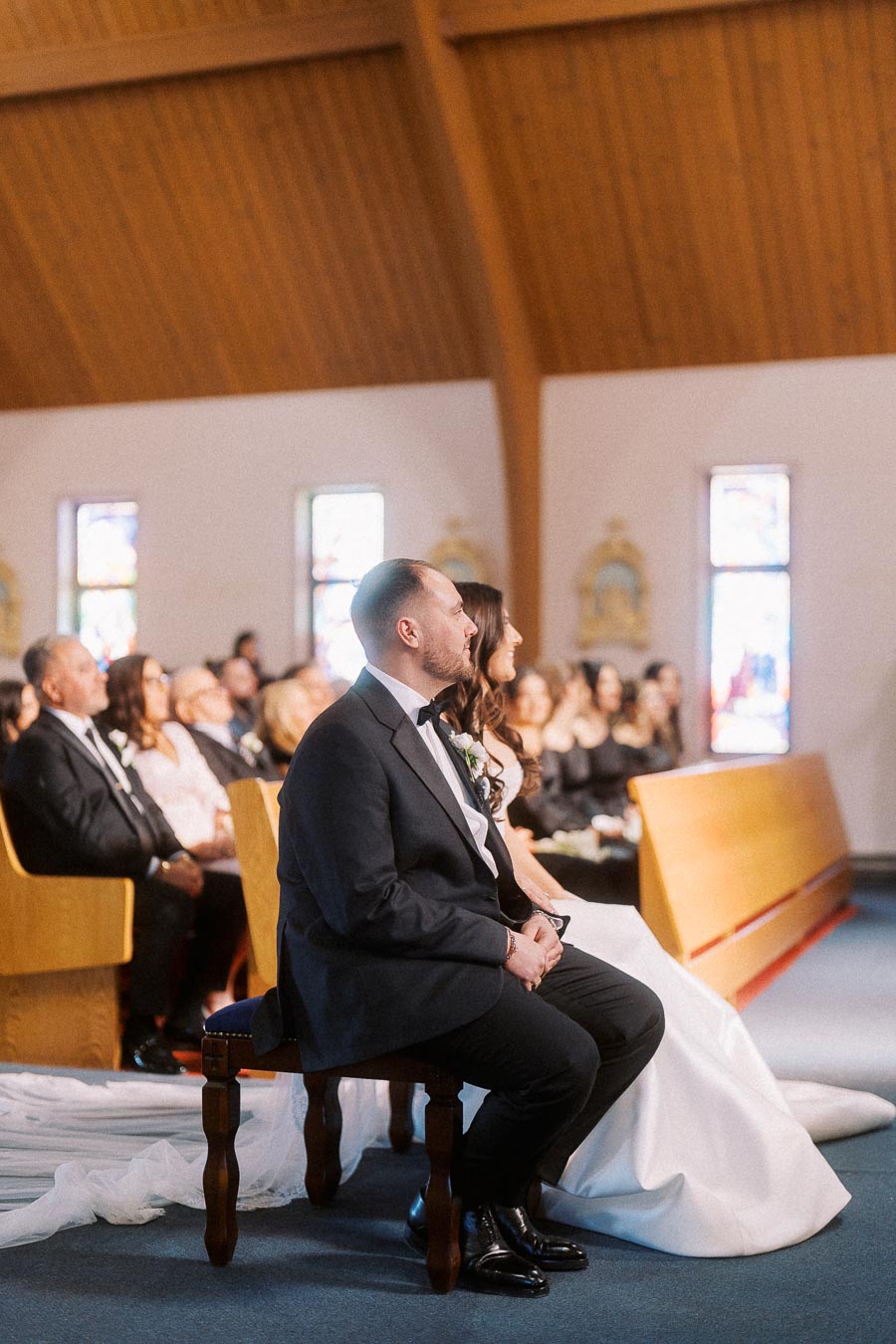 Wedding ceremony inside a church with bride and groom seated in the front row, surrounded by guests in traditional attire, under wooden vaulted ceiling and stained glass windows.
