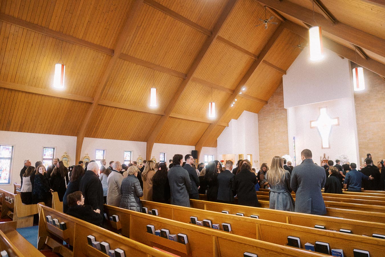 Congregation gathered inside a church interior with wooden vaulted ceiling, featuring a cross illuminated at the altar, and attendees in formal attire standing between the pews.