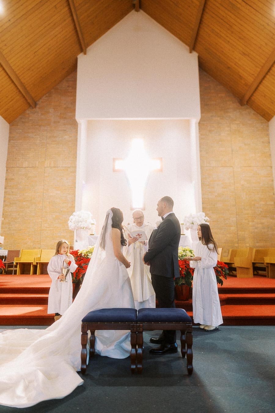 Bride and groom stand at the altar during a traditional church wedding ceremony, accompanied by a priest and two young attendants in white robes.