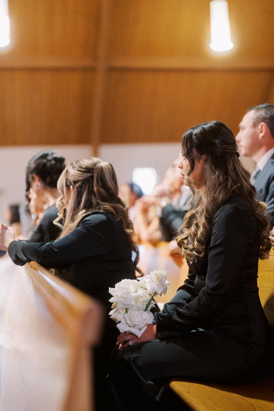 Group of people sitting in a church pew during a ceremony, with a focus on a woman holding white roses, suggesting a formal occasion or celebration.