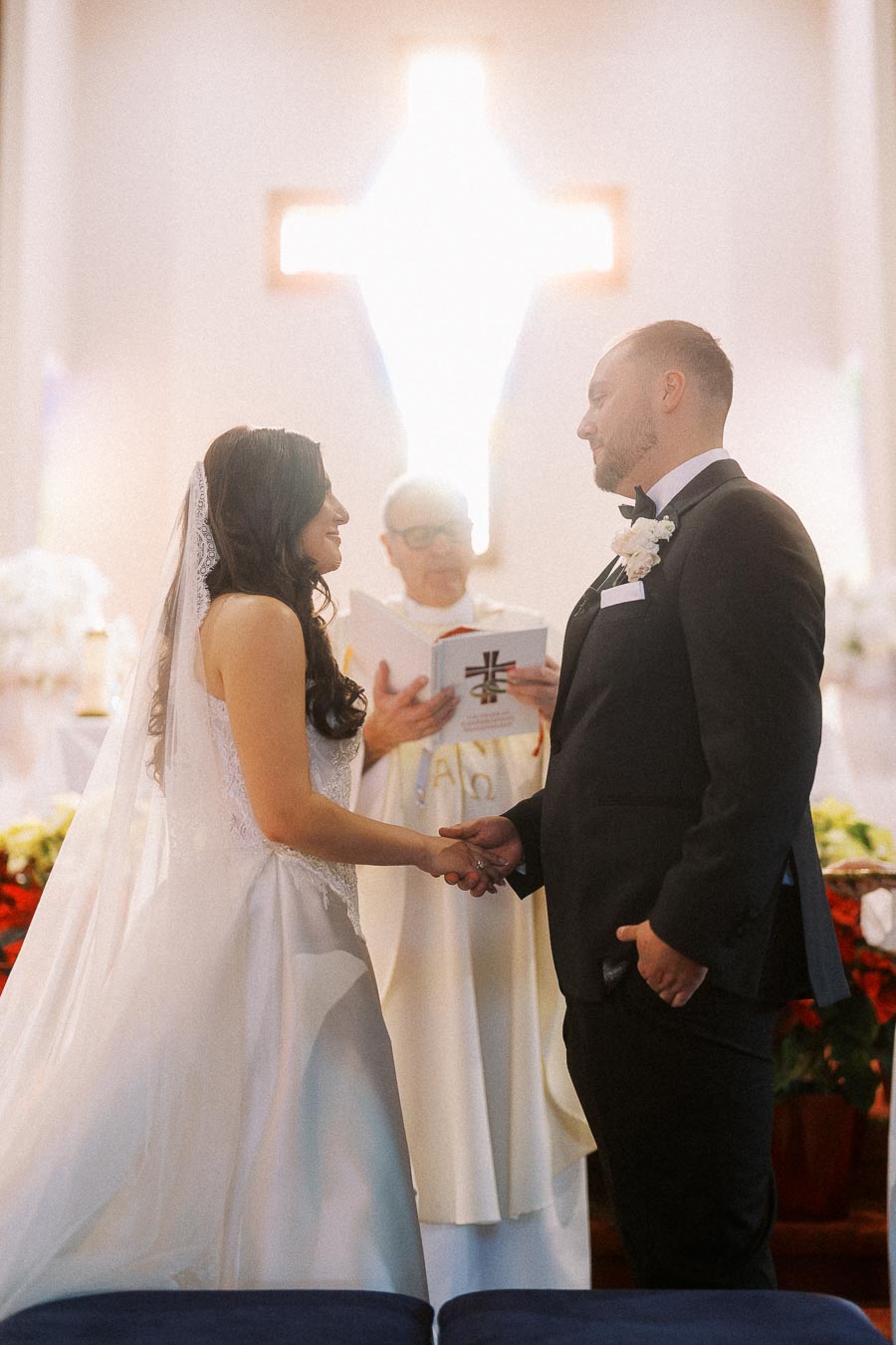 Bride and groom holding hands during their wedding ceremony in a church, with a priest officiating and a glowing cross in the background.