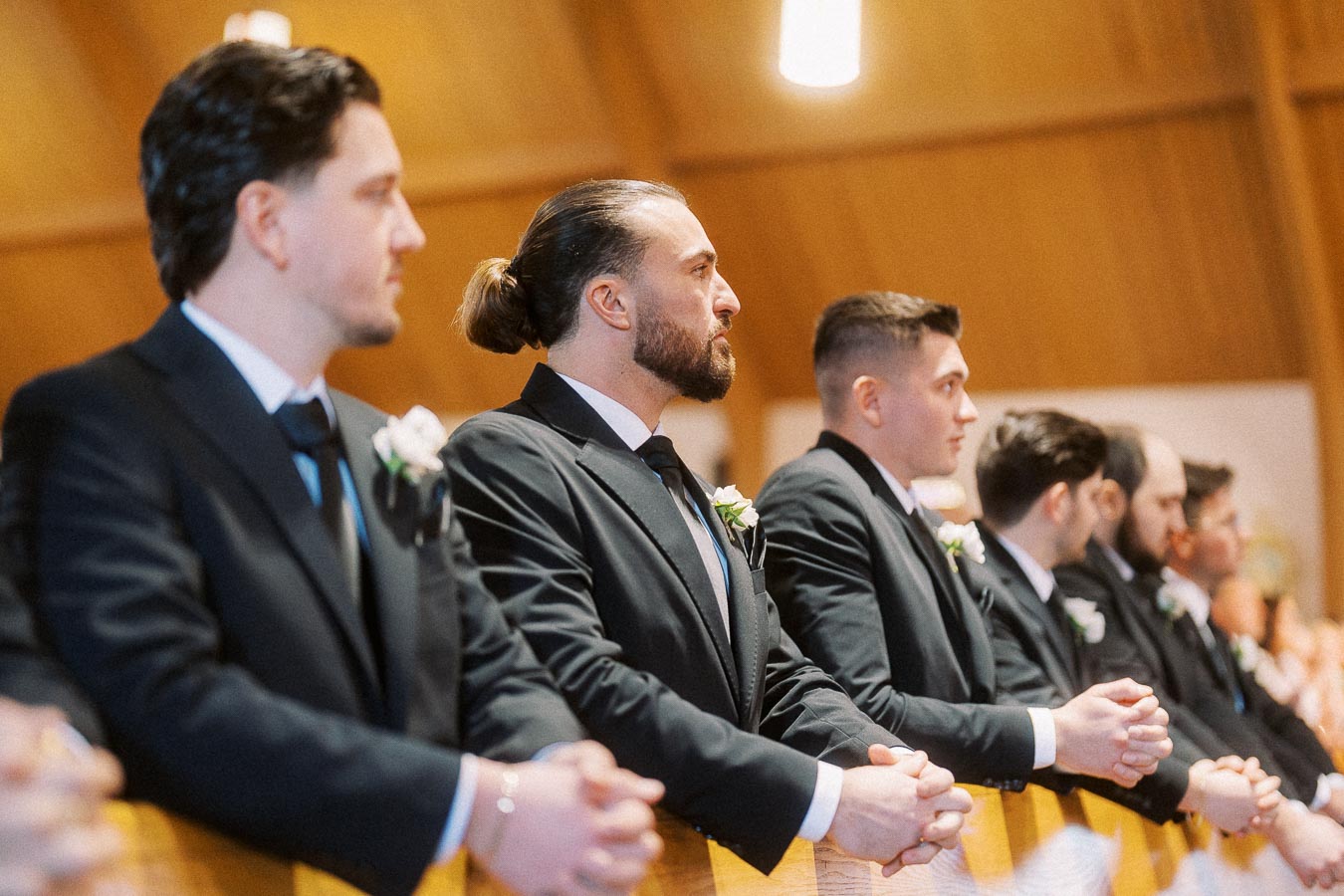 A row of groomsmen in black suits with white boutonnieres stand in a church, attentively watching a wedding ceremony.