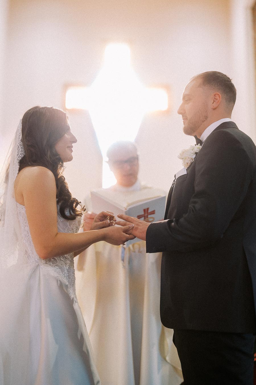 Bride and groom exchanging rings during a church wedding ceremony with a priest in the background.