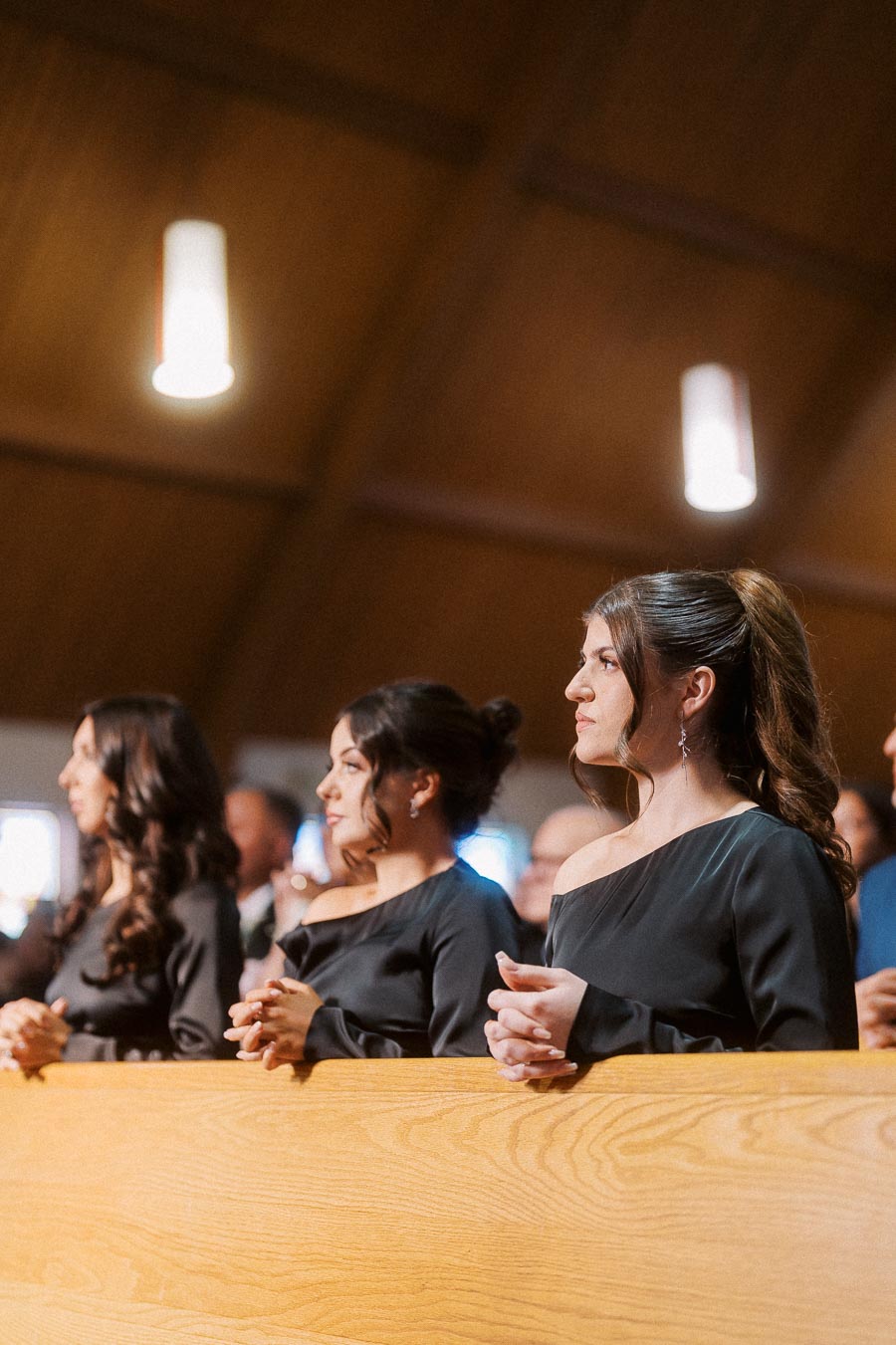 A group of women in formal black dresses standing attentively in a warmly lit church setting.