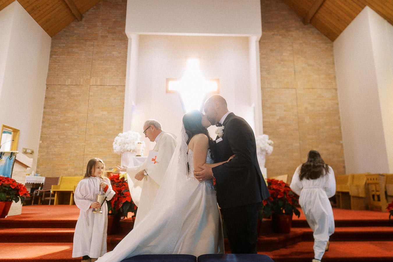 A bride and groom share a kiss at the altar during a church wedding ceremony, with a priest and a young child in white robes holding a silver chalice nearby, beneath a large illuminated cross.