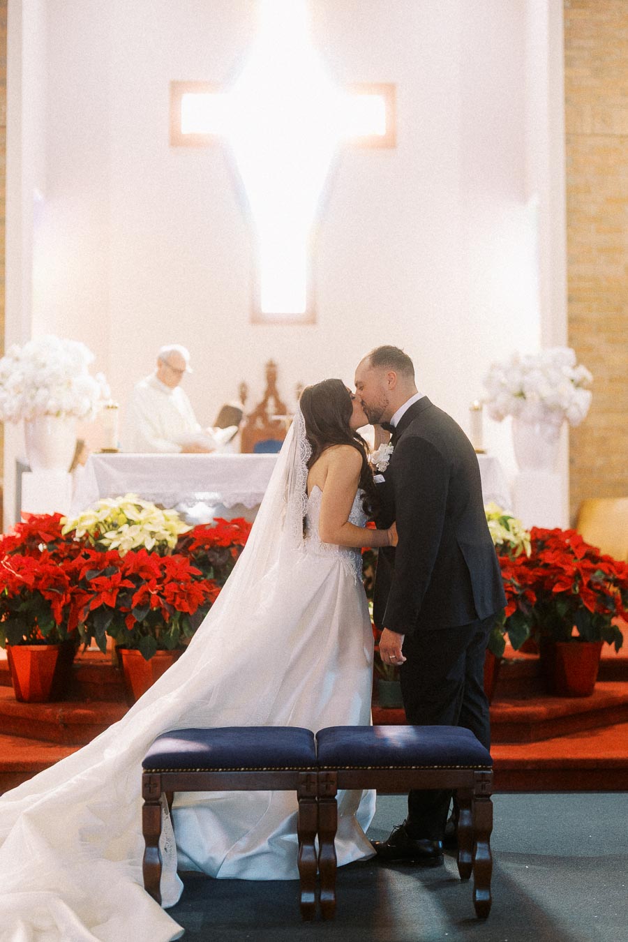 Bride and groom sharing a kiss in a beautifully decorated church with vibrant red and white floral arrangements, under a prominent illuminated cross, during their wedding ceremony.