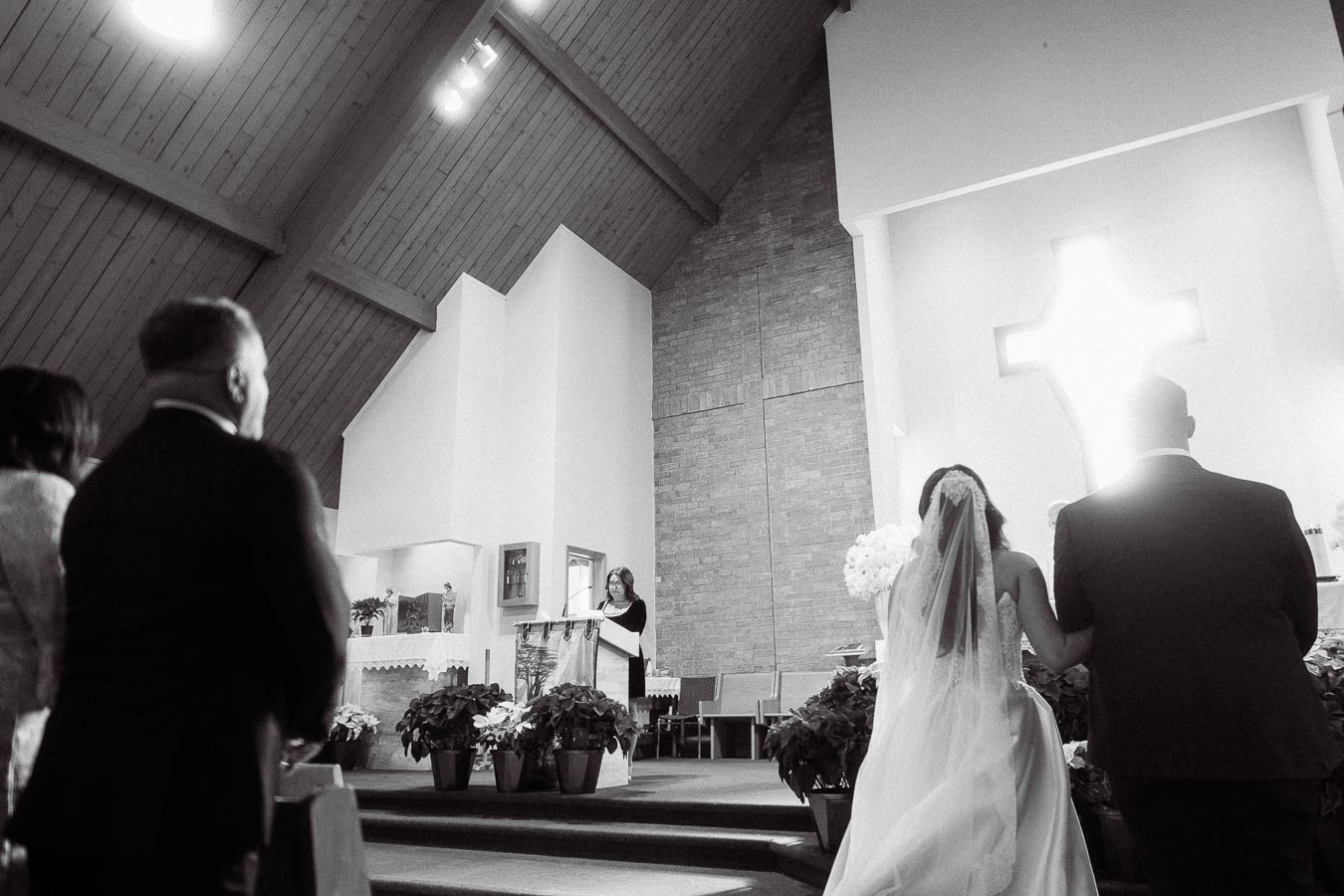 Black and white photo of a couple standing at the altar during a wedding ceremony in a church, with a large illuminated cross in the backdrop. The pews are lined with potted plants and a person reading at a podium is visible on the left.