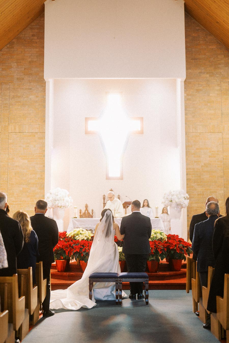 Bride and groom kneeling at the altar during a church wedding ceremony, surrounded by guests, with a priest officiating and a large illuminated cross in the background.
