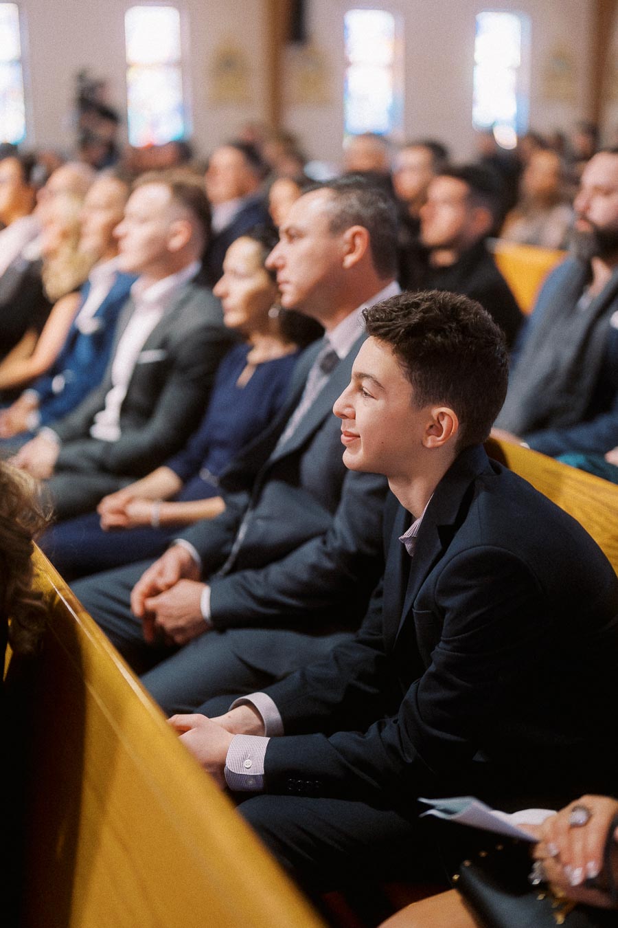 Audience attentively listening during a formal event in a church setting, featuring young and older attendees dressed in formal attire, seated in wooden pews.