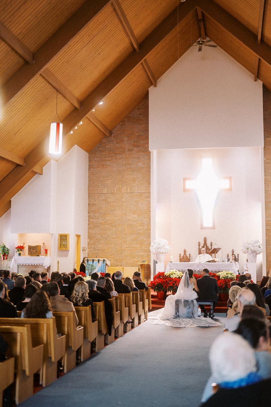 Interior of a church during a wedding ceremony with a bride and groom sitting at the altar, surrounded by guests in pews, and illuminated by natural light coming through a cross-shaped window.