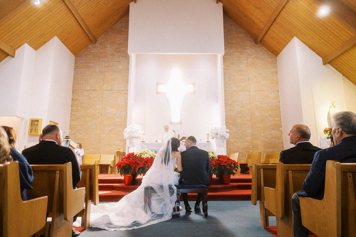Wedding ceremony in a church with a bride and groom kneeling before a priest at the altar, adorned with flowers and a glowing cross, surrounded by seated guests.