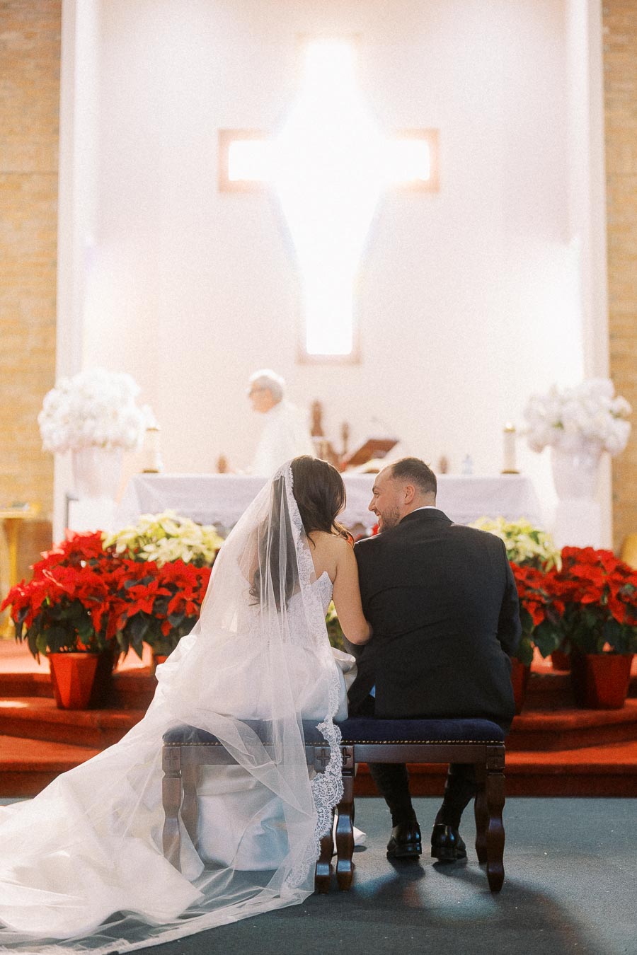 Bride and groom kneeling in church during wedding ceremony, with altar decorated with red and white poinsettias, large illuminated cross in background.
