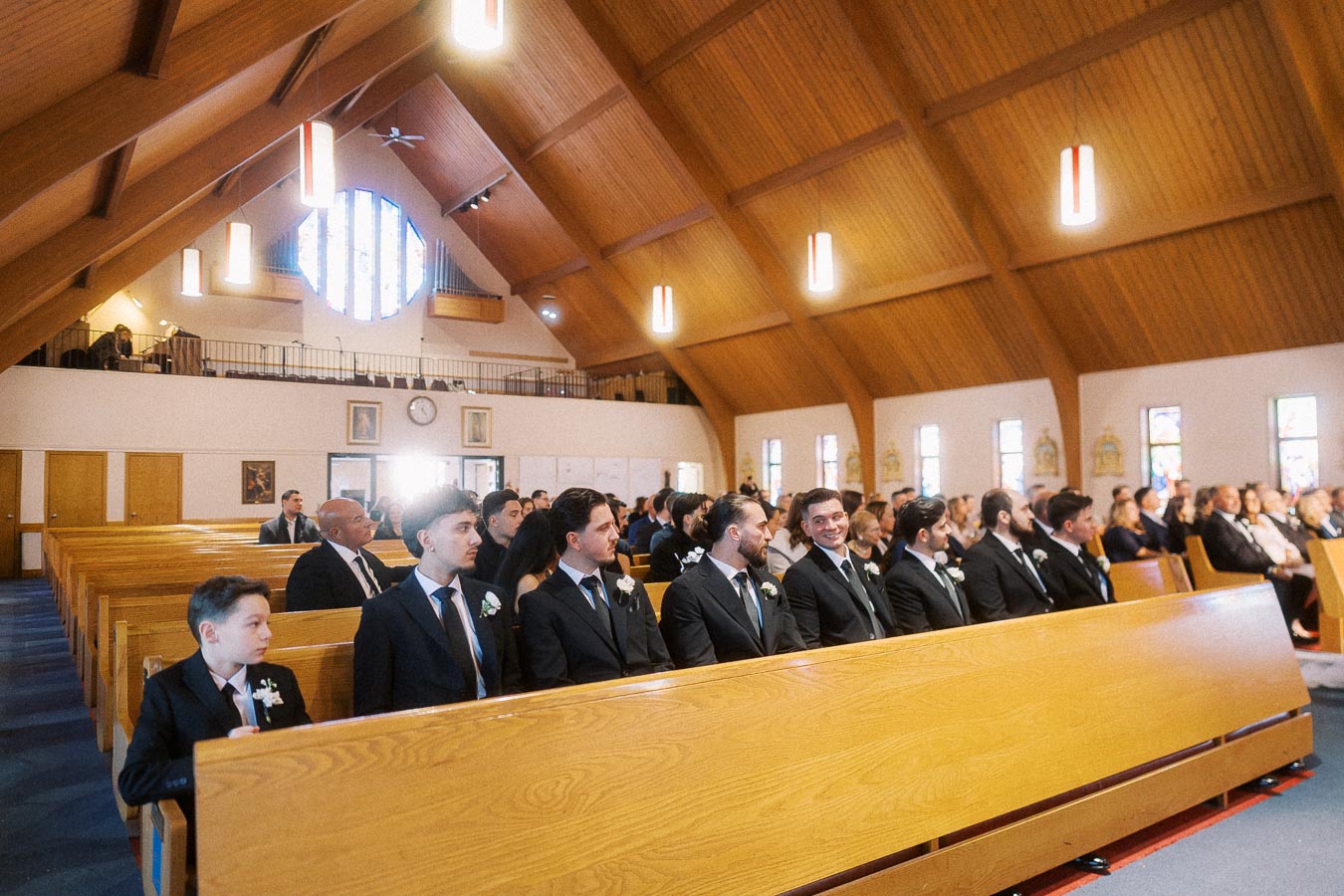 A group of people inside a beautifully lit church with wooden pews and a vaulted ceiling, dressed in formal attire, attentively awaiting a ceremony.