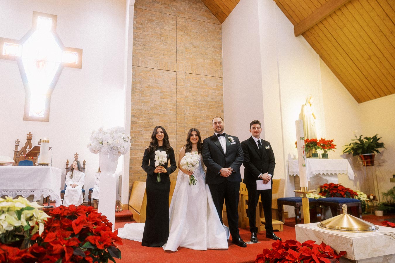 Wedding ceremony in a church with a bride and groom standing alongside their attendants. The bride is in a white gown, holding a bouquet, and the groom is in a black tuxedo. The attendants are dressed formally, with floral arrangements and red poinsettias in the foreground, and a cross and wooden ceiling in the background.