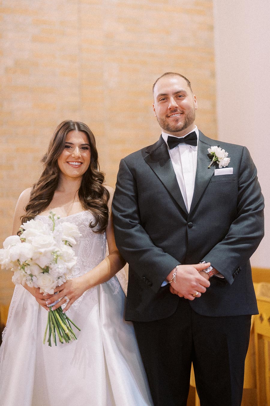 A bride in a white wedding gown holding a bouquet of white flowers stands next to a groom in a black tuxedo with a boutonniere, both smiling, inside a warmly lit venue.