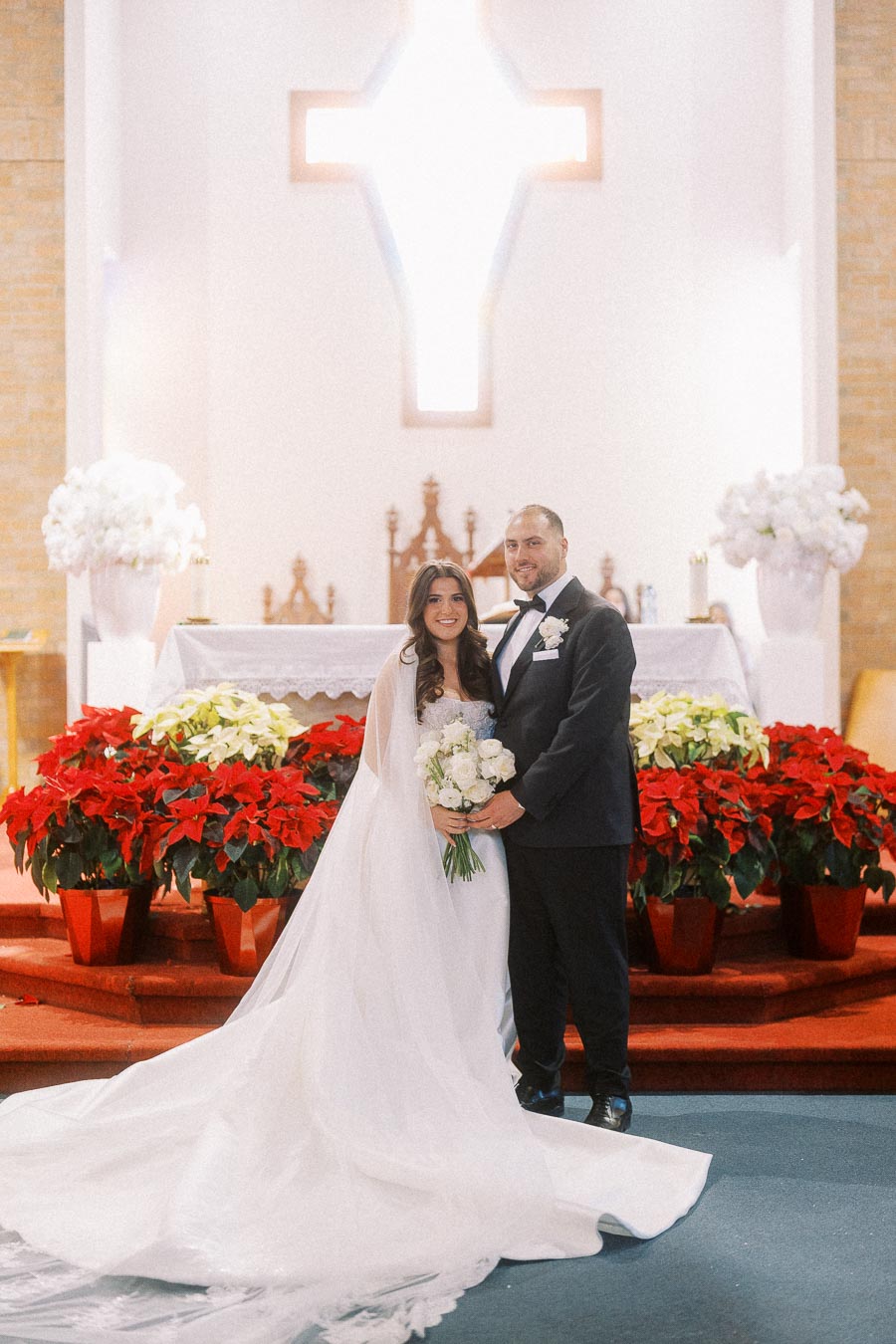 Bride and groom posing in a church adorned with red and white flowers, standing in front of an altar and illuminated cross, capturing a beautiful wedding moment.