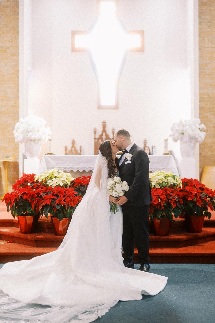 Bride and groom sharing a kiss at the altar in a beautifully decorated church, with a bright cross in the background and vibrant red poinsettias adding a festive touch to the scene.