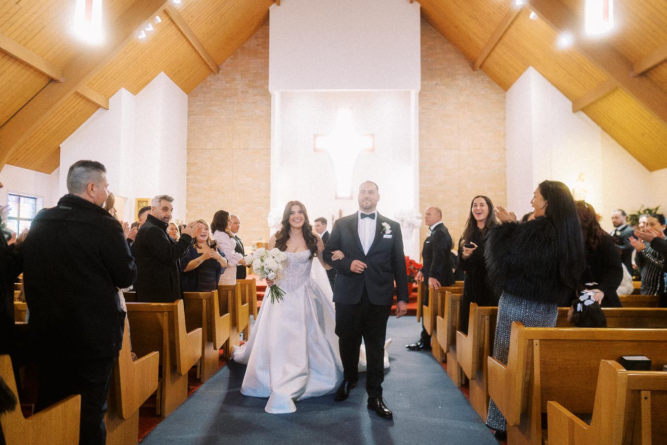 Bride and groom walking down the aisle in a church surrounded by smiling and applauding guests on their wedding day.