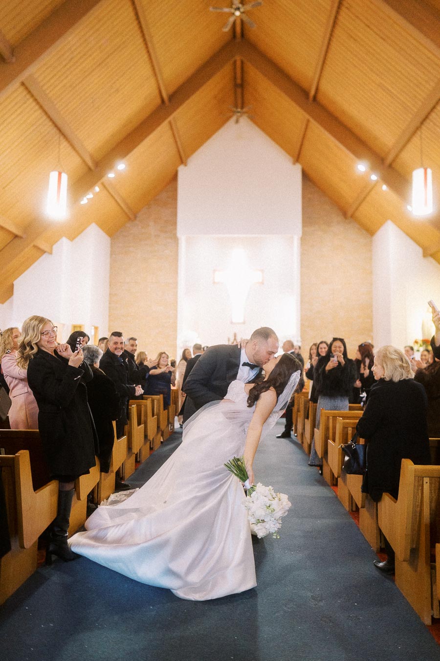 Bride and groom share a romantic kiss in a church aisle surrounded by joyful guests, with the bride holding a bouquet and the backdrop of a cross, capturing a special wedding moment.