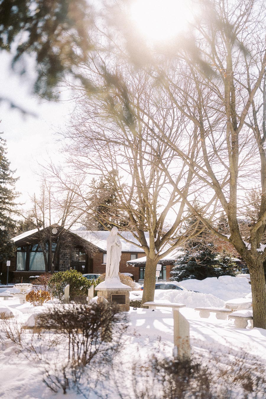 Winter scene of a serene snowy garden featuring a white statue amidst bare trees, bright sunlight filtering through branches, and a brick building in the background.