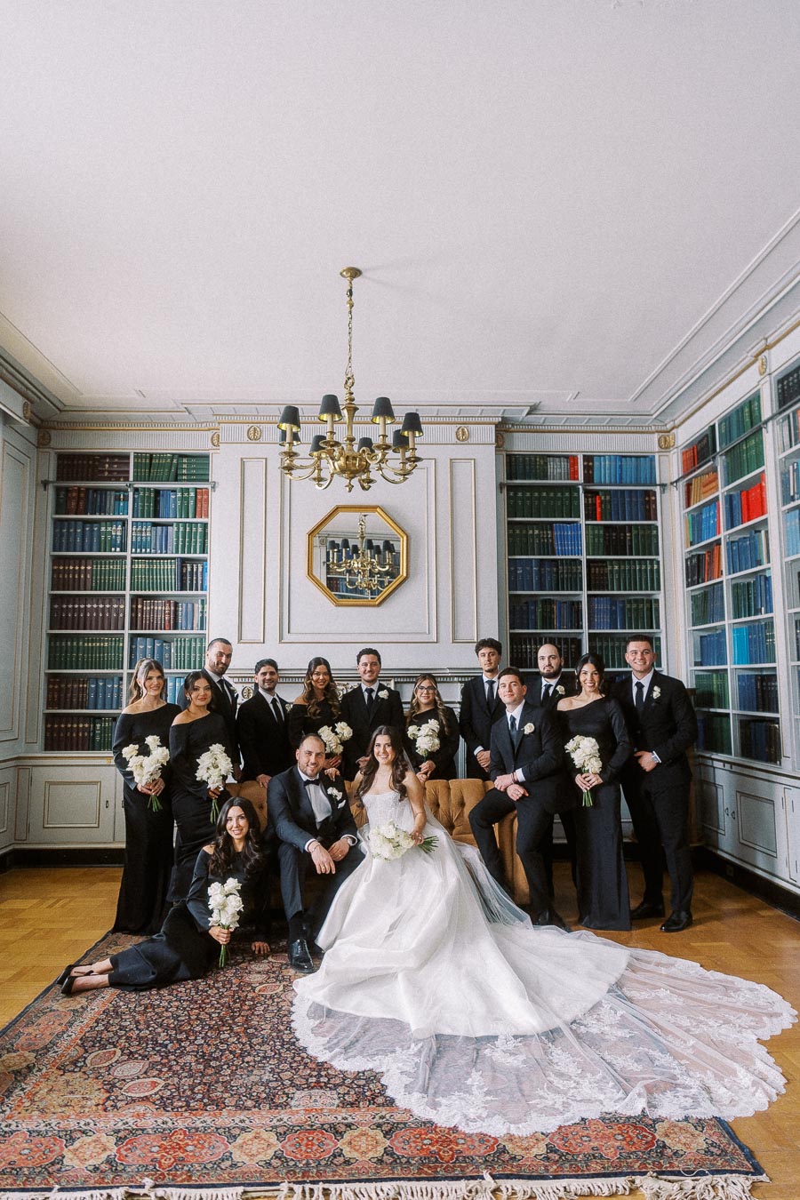 Elegant wedding party portrait in a sophisticated library setting, featuring the bride in a stunning white gown and the groom in a classic tuxedo, surrounded by bridesmaids and groomsmen in formal black attire, all holding white bouquets.