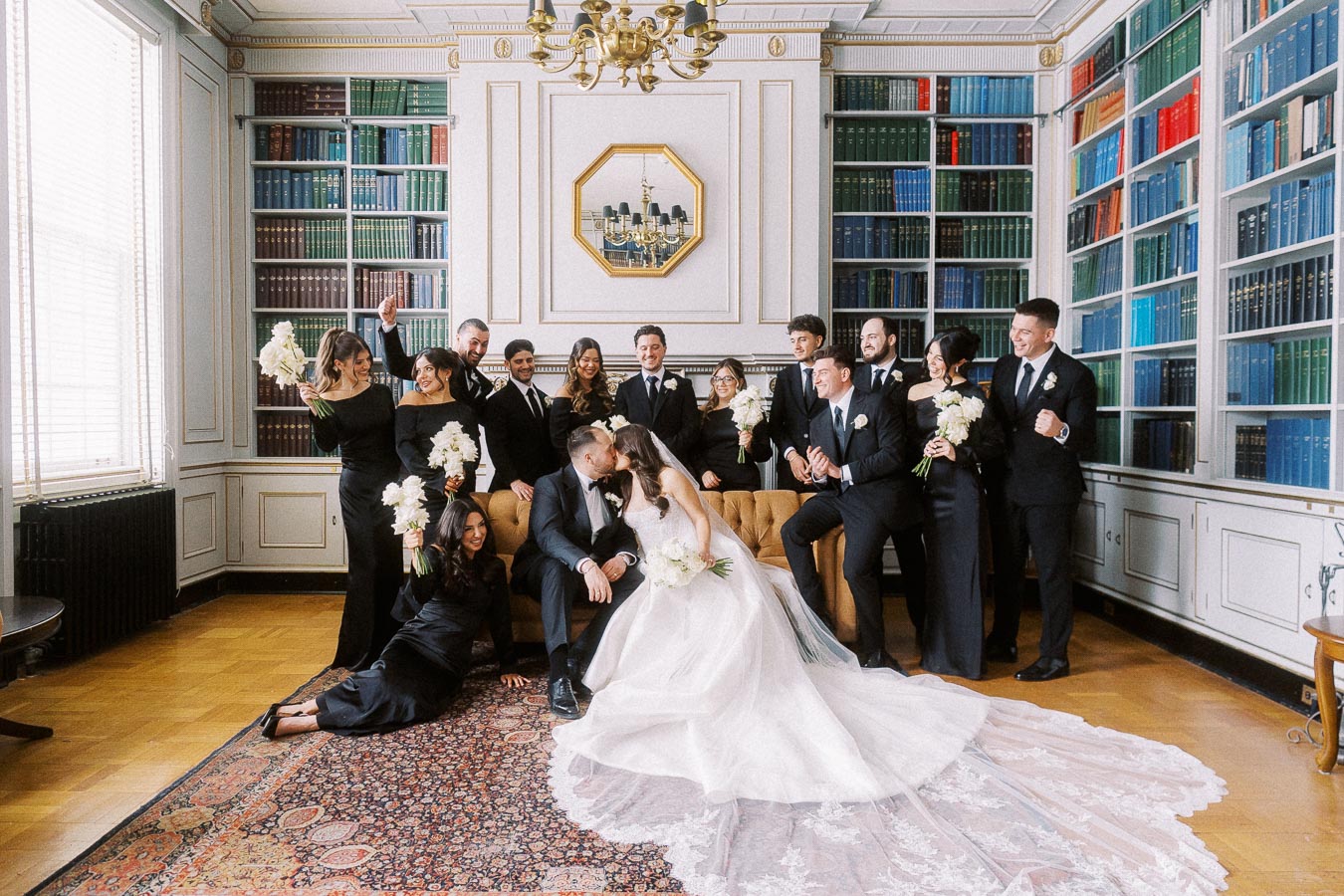 Elegant wedding party in a library with bride in a voluminous gown and groom in black tuxedo, surrounded by bridesmaids in black dresses and groomsmen in suits, celebrating joyfully in front of bookshelves and chandelier.