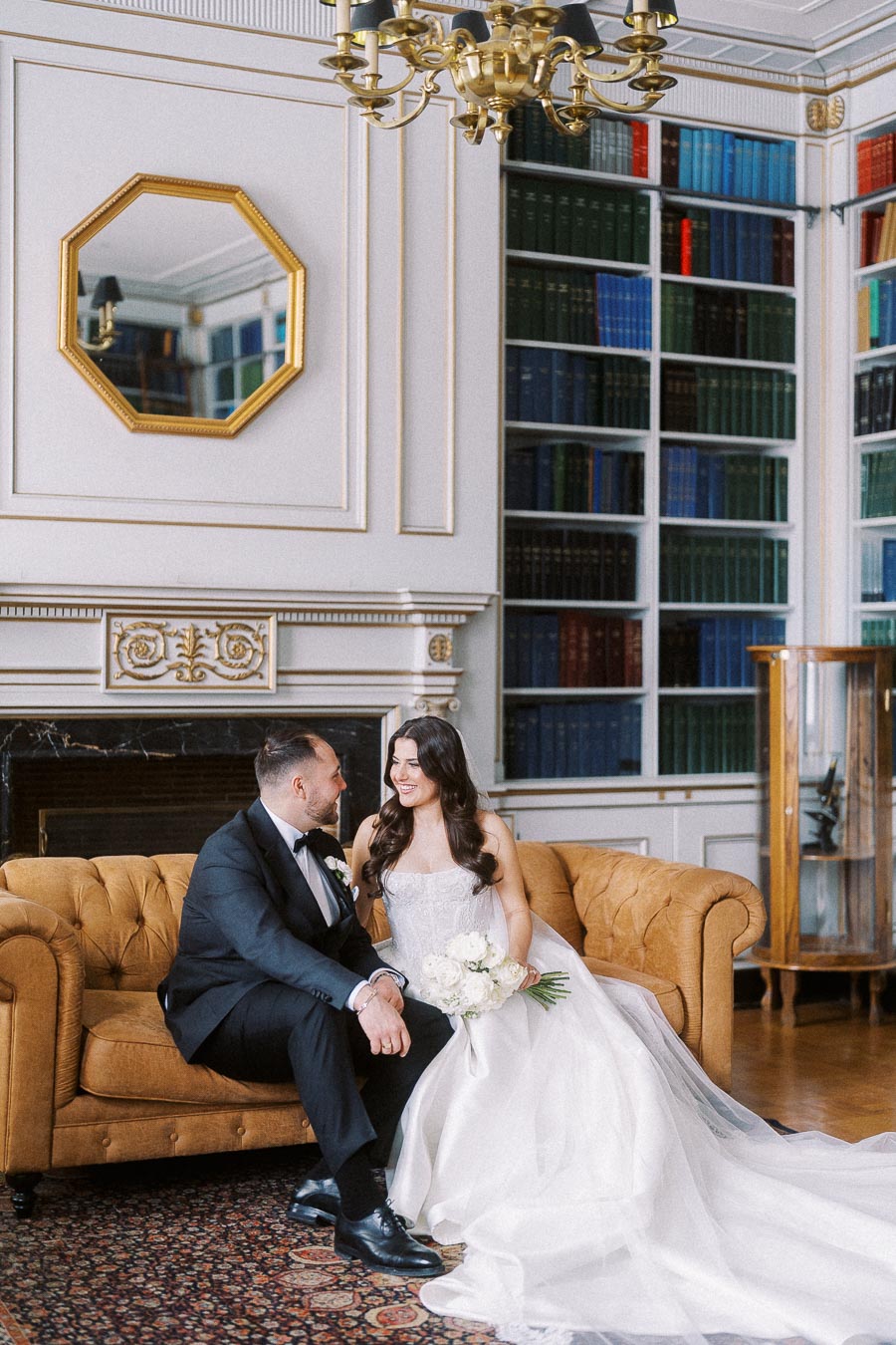 A bride and groom sit on a luxurious tan sofa in an elegant wedding photoshoot, surrounded by antique bookshelves and ornate decor in a classic library setting.