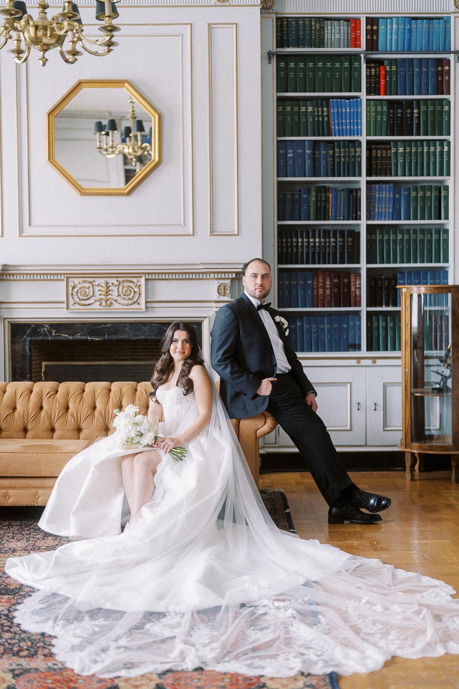 Elegant bride in white dress with lace details holding bouquet, seated on a vintage sofa next to groom in black tuxedo, posed in a sophisticated library setting with bookshelves and ornate decor.