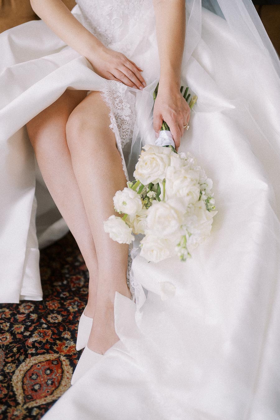 Bride in an elegant white lace wedding dress, gently holding a bouquet of white roses, seated on a patterned carpet, capturing a moment of serene elegance.
