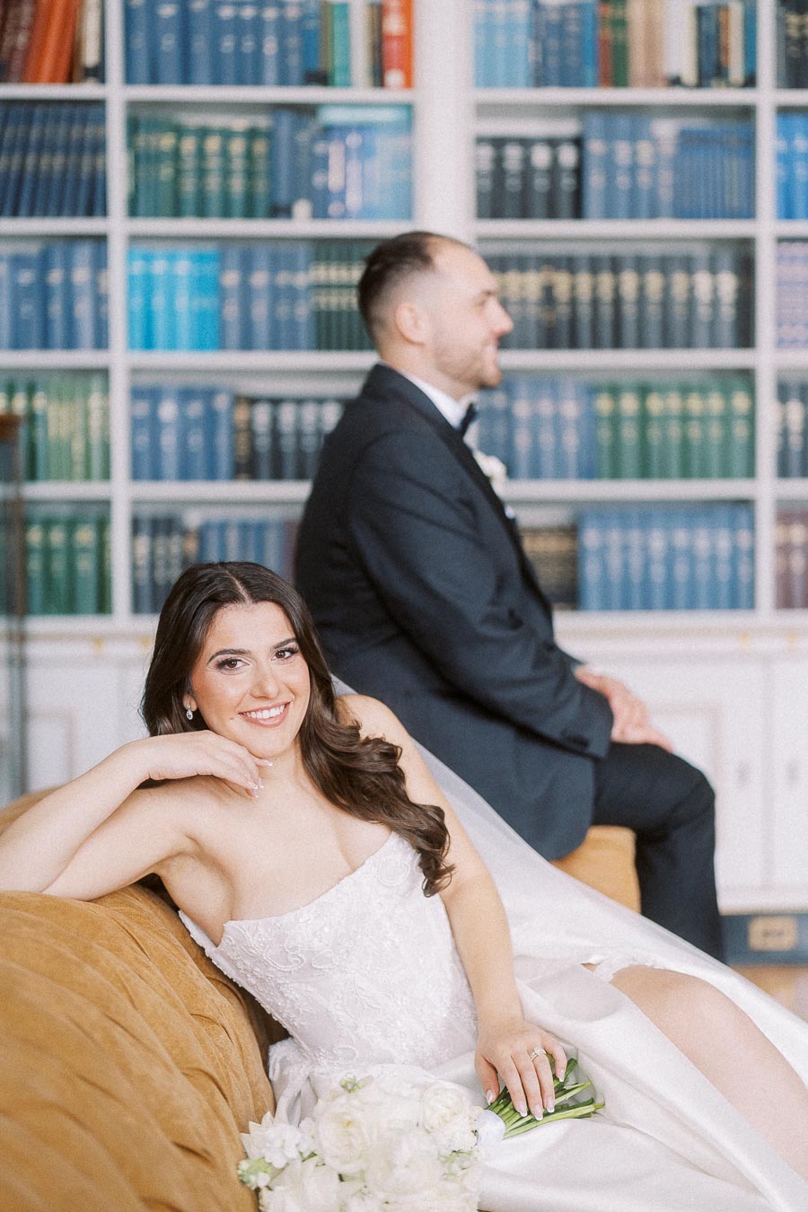 Bride in elegant white gown posing on a vintage couch with groom in background, set against a backdrop of colorful bookshelves.