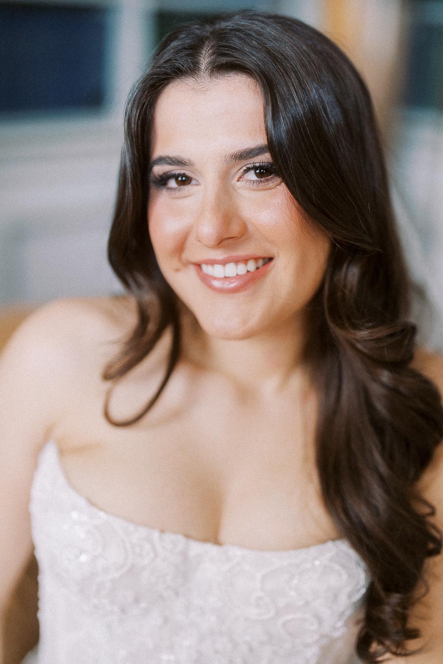 Smiling bride with long brown hair wearing an elegant lace wedding dress indoors.