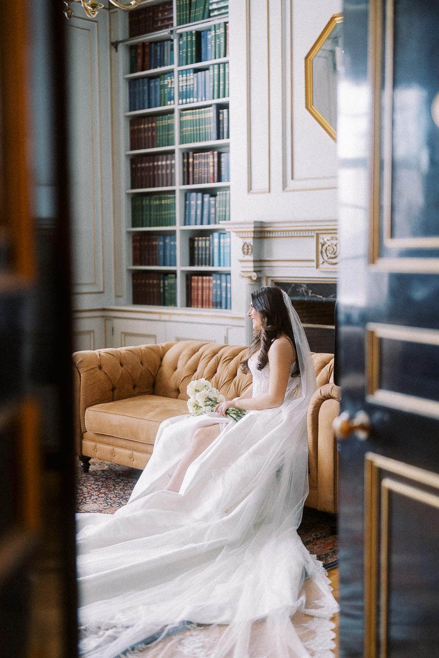 Bridal portrait in elegant library setting, featuring a seated bride in a flowing white gown and veil, holding a bouquet of white flowers, with a classic leather couch and a backdrop of filled bookshelves.