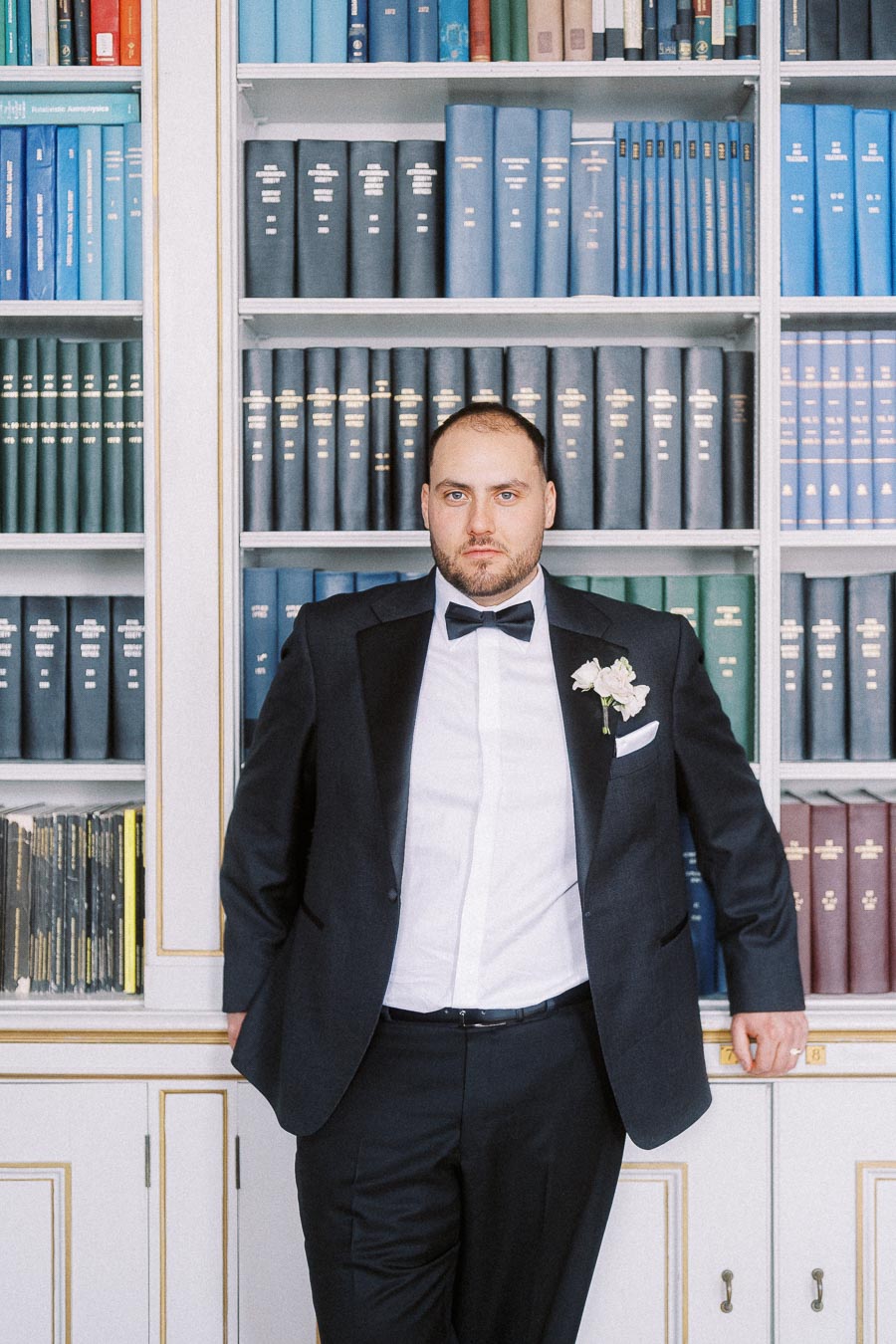 Man in a tuxedo with a bow tie and floral boutonniere standing confidently in front of a bookshelf filled with colorful books.