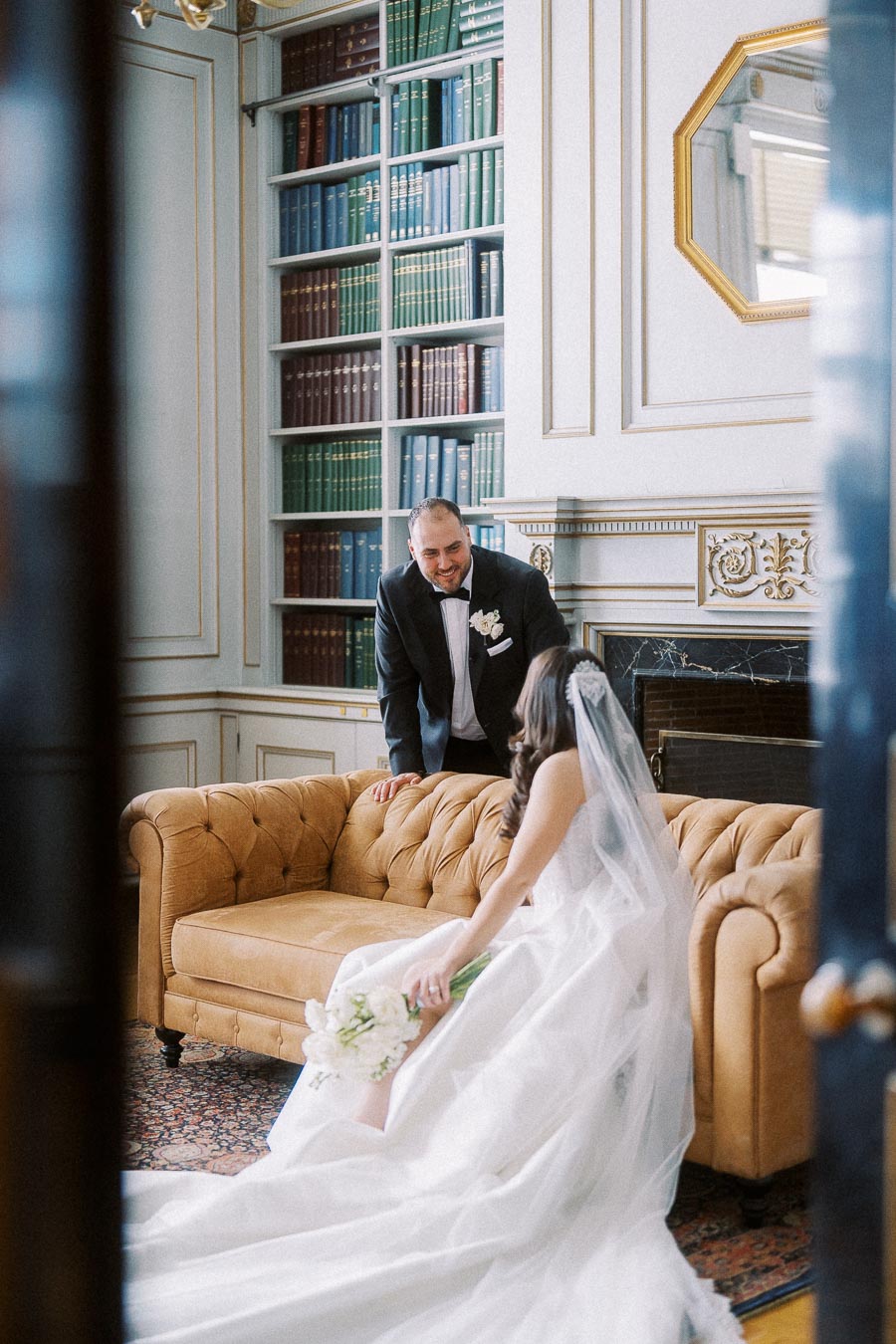 Elegant bride and groom in a sophisticated library setting, with the bride seated on a vintage couch in a flowing white gown and veil, and the groom leaning in, dressed in a classic black tuxedo.