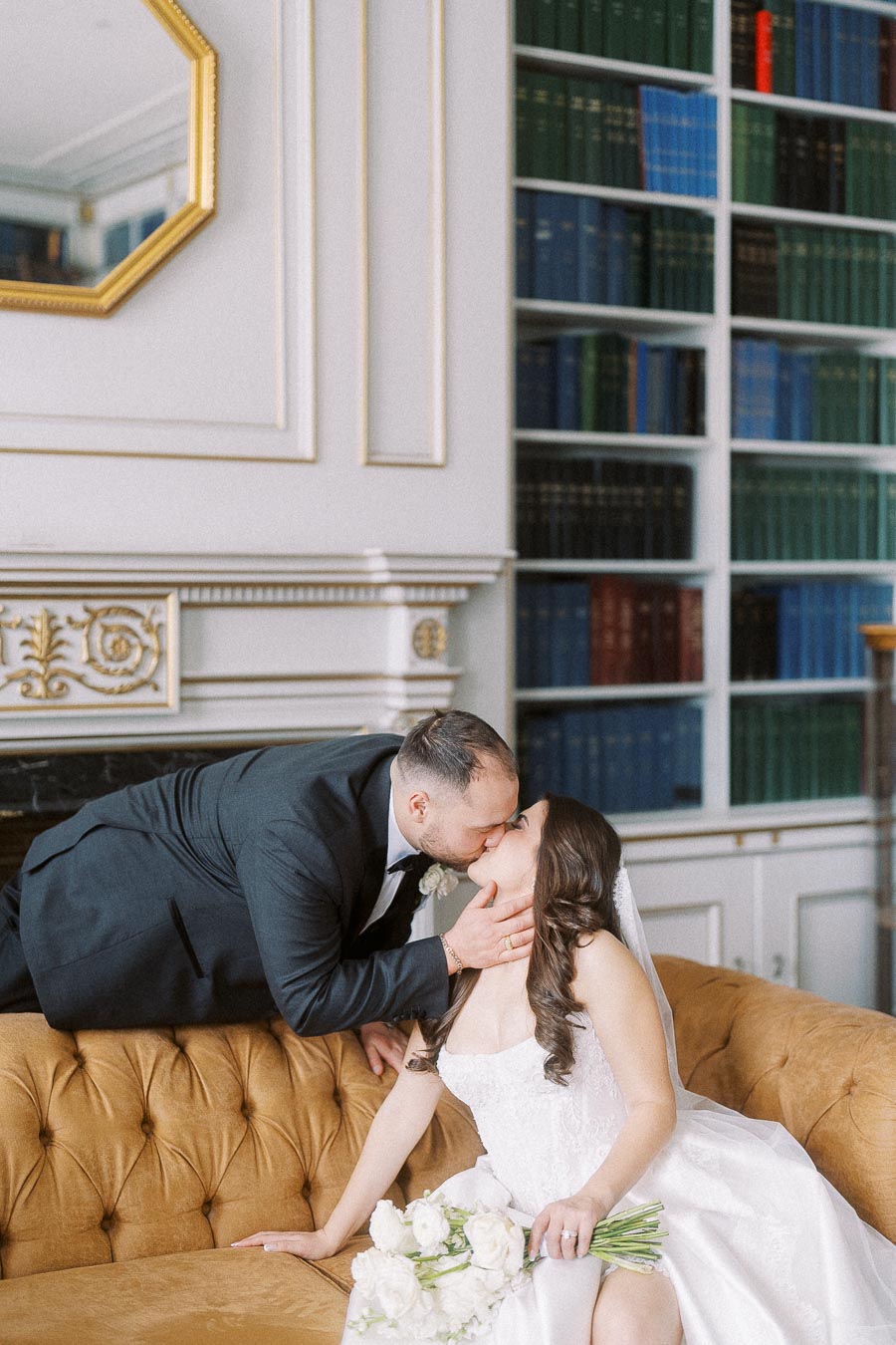 Romantic wedding moment with newlywed couple kissing on a vintage sofa, bride holding white flower bouquet in elegant library setting.