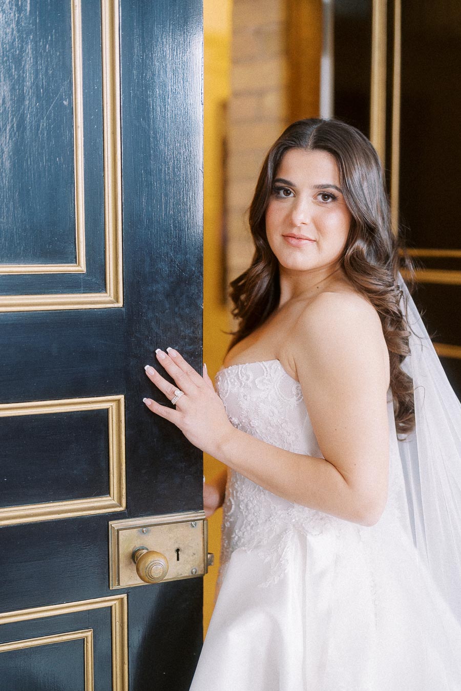 Bride in elegant white wedding gown standing by ornate door, showcasing classic beauty with curled hair and veil, perfect for modern wedding inspiration.