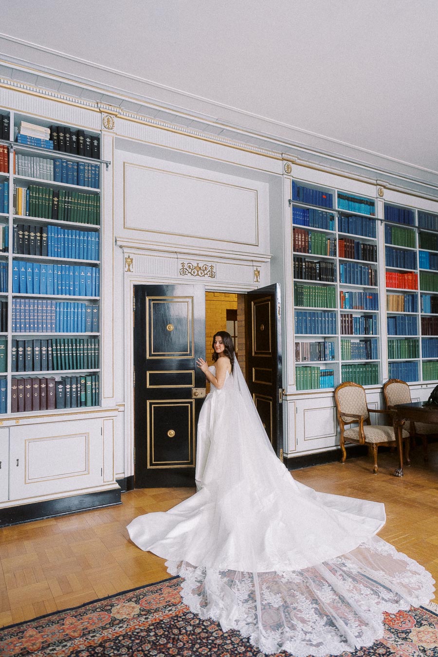 Bride in elegant white wedding dress with long lace train, standing in a luxurious library with ornate bookshelves and classic wooden door.