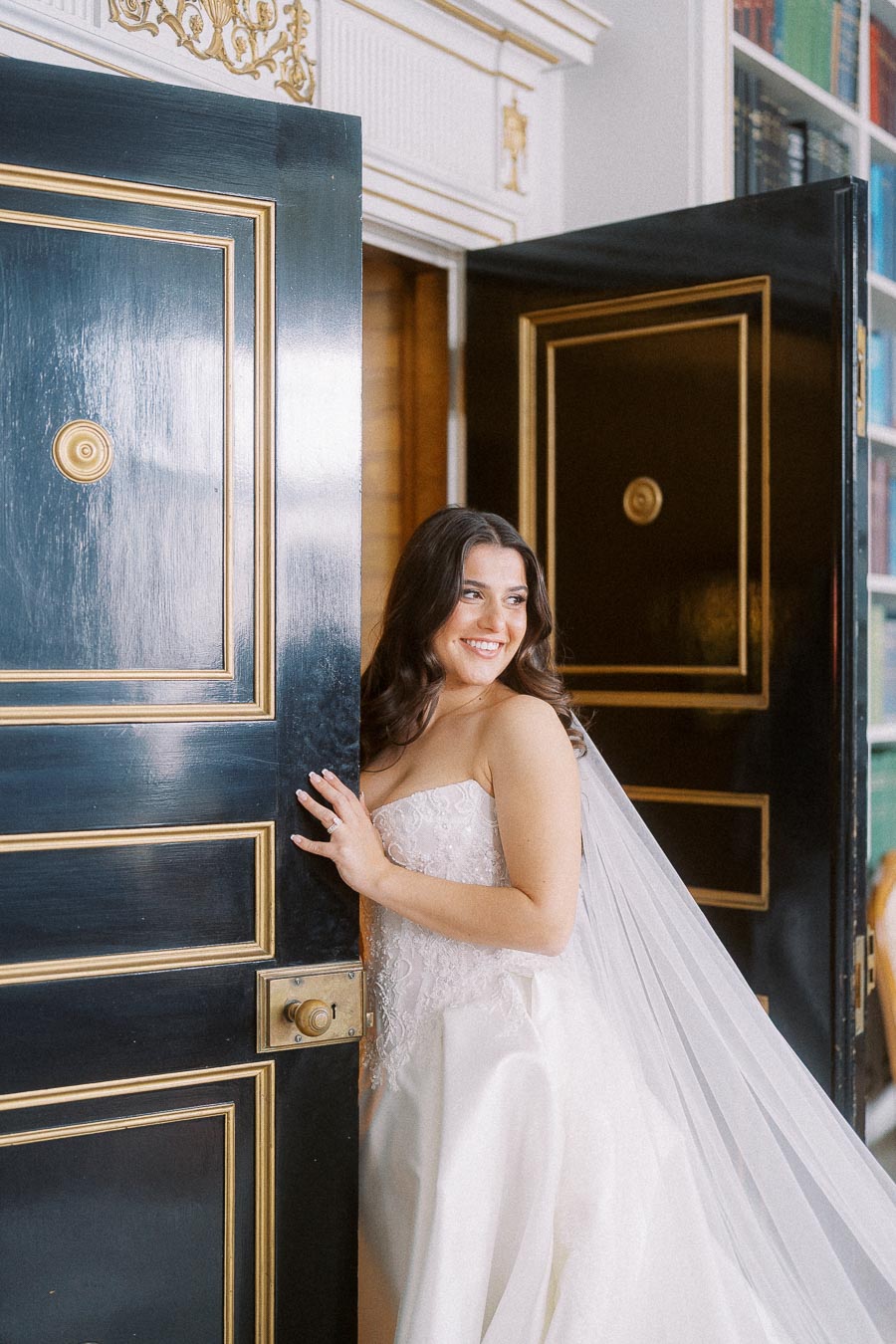 A smiling bride in a strapless wedding gown peeks through an elegant doorway, with gold detailing on the woodwork, capturing a moment of joy and anticipation on her wedding day.