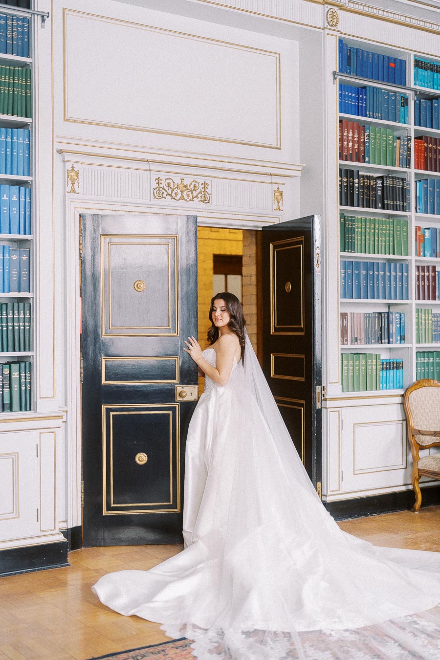 Bride in elegant wedding gown and long veil standing in a sophisticated library with bookshelves, ornate wooden door, and classic decor.