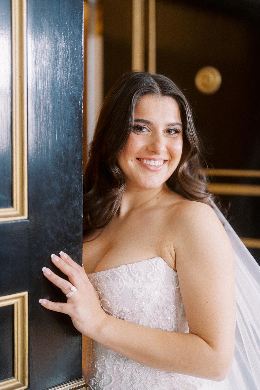 Smiling bride in elegant lace wedding dress poses by black and gold door.