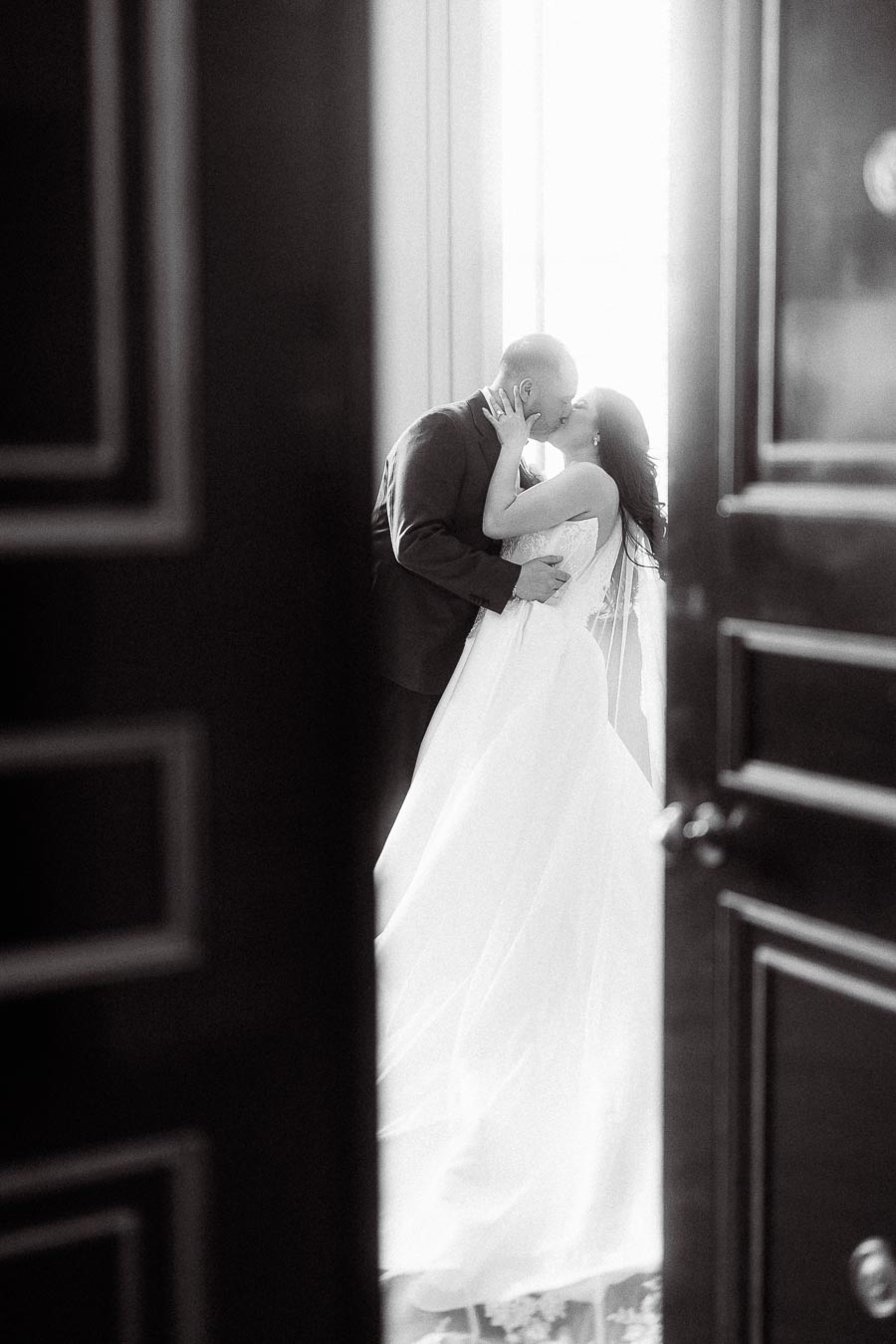 Black and white image of a romantic moment as a bride and groom share a kiss near a window, captured through the open doorway.